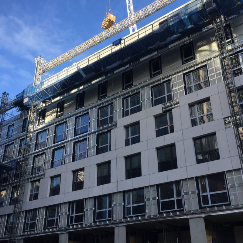 Image of the Travelodge Aldgate building during construction, partway through the installation of lightweight cladding and the full facade. The building's exterior features partially installed panels, exposing the subframe and insulation layers, with visible scaffolding and construction materials on-site.