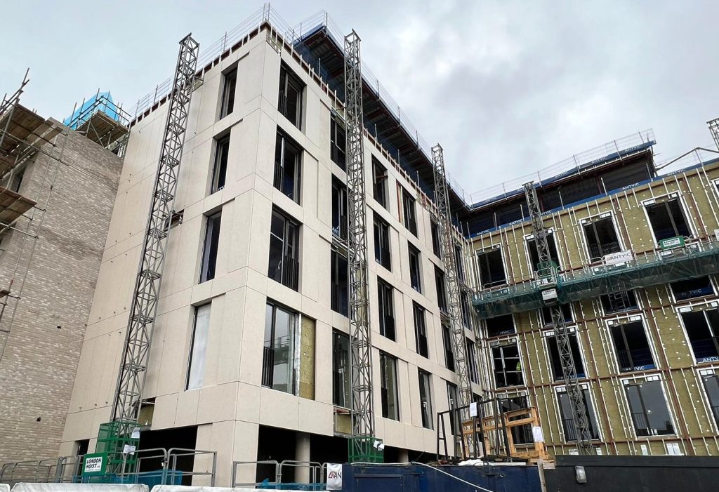 Image of the Hazel Court building during construction, partway through the installation of porcelain cladding and the full facade. The building's exterior features partially installed panels, exposing the subframe and insulation layers, with visible scaffolding and construction materials on-site.